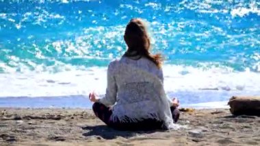 Young woman sitting cross-leged near the sea and streching in windy weather