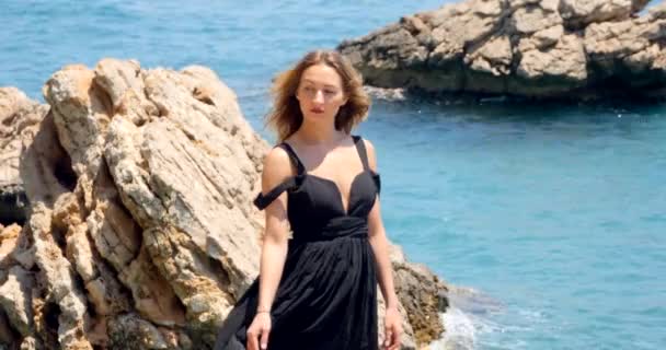 Νεαρή όμορφη γυναίκα σε μαύρο αποκόμματα κομψό φόρεμα κάθεται κοντά στη θάλασσα — Αρχείο Βίντεο
