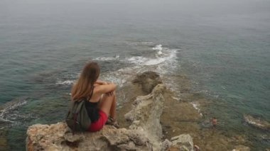Sırt çantası ile kadın turist açık gökyüzü ve sakin deniz arka plan üzerinde bir kaya üzerinde oturan