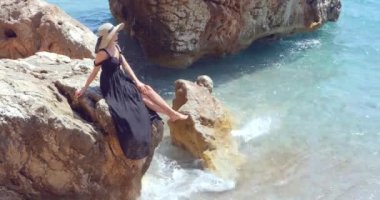 Young pretty woman in black elegant dress sitting on the rocks near the sea