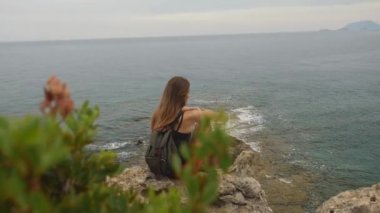 Sırt çantası ile kadın turist açık gökyüzü ve sakin deniz arka plan üzerinde bir kaya üzerinde oturan
