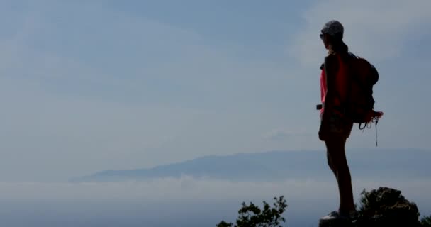 Young woman with backpack standing on the edge of mountain above the clouds — Stock Video