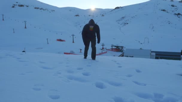 Молодой человек в темной одежде, падающий измотанный на снежной горе — стоковое видео
