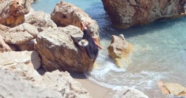 Young pretty woman in black elegant dress sitting on the rocks near the sea