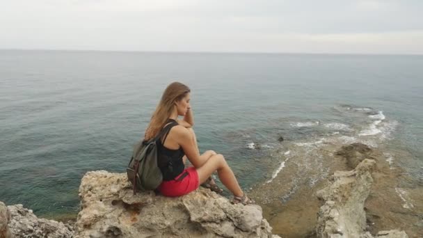 Женщина туристка с рюкзаком сидит на скале на ясном небе и спокойном морском фоне — стоковое видео