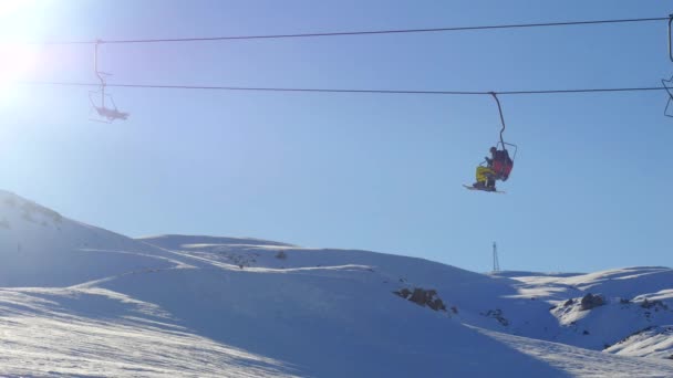 Vista do elevador de esqui em movimento com pessoas e atletas snowboard de montanha nevada no fundo — Vídeo de Stock