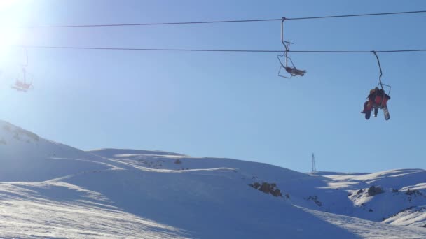 Uitzicht op de bewegende skilift met mensen en sporters snowboarden van besneeuwde berg op de achtergrond — Stockvideo