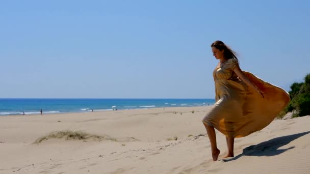 Joven mujer hermosa en vestido largo dorado de pie sobre dunas de arena cerca del mar . — Vídeos de Stock