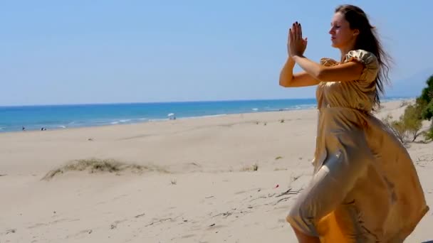 Νέα όμορφη γυναίκα σε χρυσό μακρύ φόρεμα στέκεται σε αμμόλοφους κοντά στη θάλασσα. — Αρχείο Βίντεο