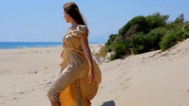 Young beautiful woman in golden long dress standing on sand dunes near the sea.