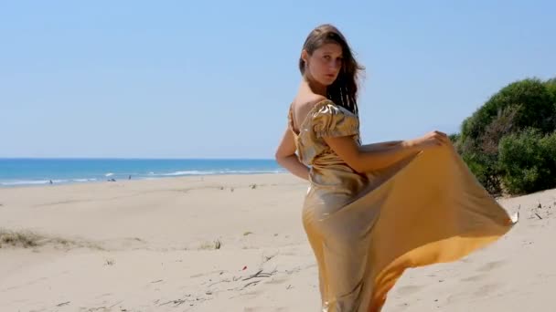 Jonge mooie vrouw in gouden lange jurk staande op zandduinen in de buurt van de zee. — Stockvideo