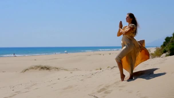 Young beautiful woman in golden long dress standing on sand dunes near the sea. — Stock Video