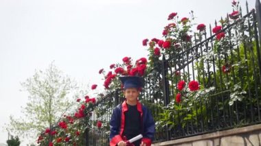 Mezuniyet elbisesi içinde mutlu beyaz çocuk, vahşi güllerle dolu taş çitin yanında duran diploması yla