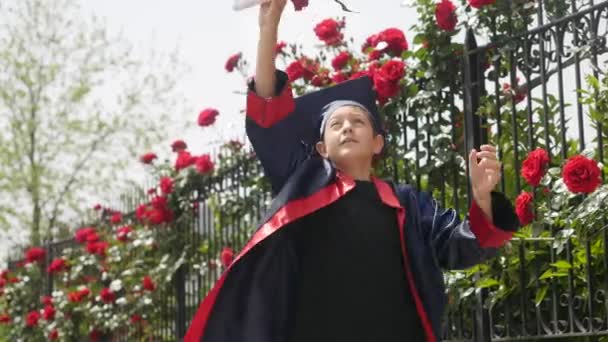 Happy kaukasiska barn i examen klänning med diplom glatt dansa nära staket full av vilda rosor — Stockvideo