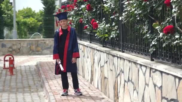 Niño caucásico feliz en vestido de graduación con diploma de pie cerca de valla de piedra llena de rosas silvestres — Vídeos de Stock