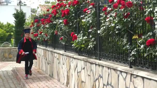Szczęśliwy kaukaski dziecko w sukni ukończenia studiów z dyplomem chodzenie wzdłuż kamiennego ogrodzenia. — Wideo stockowe