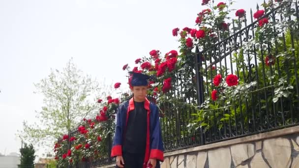 Щаслива біла дитина у випускному платті з дипломом радісно танцює біля паркану, повного диких троянд — стокове відео