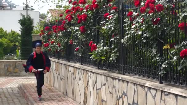 Felice bambino caucasico in abito da laurea con diploma passeggiando lungo la recinzione di pietra . — Video Stock