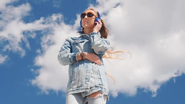 Young woman in sunglasses and jeans wear listening music with headphones outdoors — 비디오