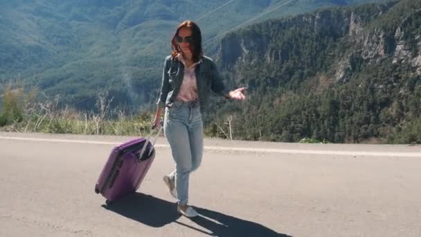 屋外の晴れた日にスーツケースヒッチハイクと田舎道の美しい若い女性 — ストック動画