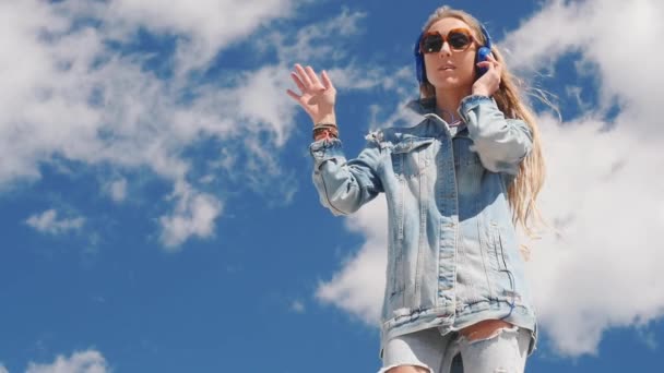 Mujer joven en gafas de sol y jeans usan música para escuchar con auriculares al aire libre — Vídeo de stock