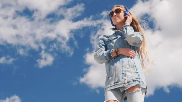 Mujer joven en gafas de sol y jeans usan música para escuchar con auriculares al aire libre — Vídeos de Stock