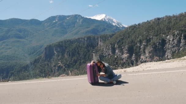 Hermosa joven sentada agotada en el camino rural con maleta haciendo autostop en un día soleado al aire libre — Vídeo de stock