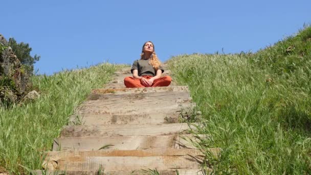 Молодая кавказская женщина сидит на деревянной лестнице и наслаждается жизнью в солнечную погоду — стоковое видео