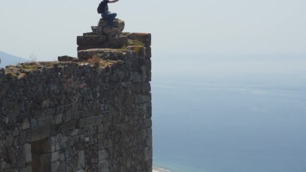 Homem turista sentado no topo da antiga parede do castelo e filmagem de vídeo no bolso câmera gimbal — Vídeo de Stock