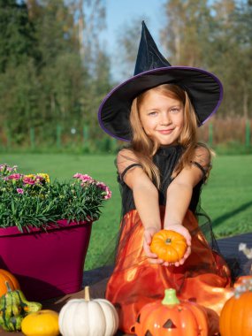 Cadı kostümlü kız tahta terasta balkabağıyla oturuyor.