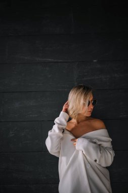 Beyaz elbiseli şık bir kadın güneş gözlüğü takarken siyah bir duvarın yanında duruyor..