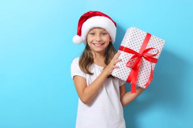 Renkli arka planda Noel şapkası takmış, elinde bir hediye tutan tatlı, neşeli bir kız.