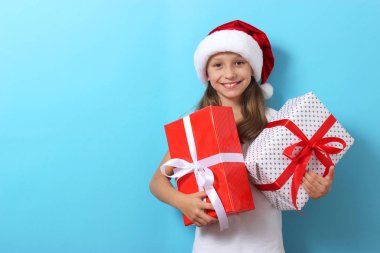 Renkli bir arka planda, Noel şapkası takmış, elinde hediyeler tutan sevimli, neşeli bir kız.