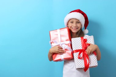 Renkli bir arka planda, Noel şapkası takmış, elinde hediyeler tutan sevimli, neşeli bir kız.