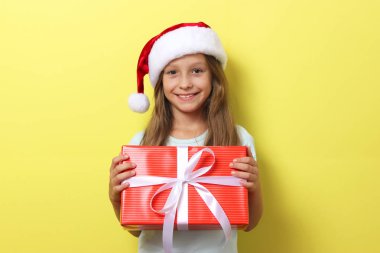 Renkli arka planda Noel şapkası takmış, elinde bir hediye tutan tatlı, neşeli bir kız.