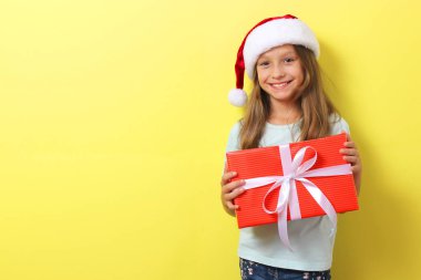 Renkli arka planda Noel şapkası takmış, elinde bir hediye tutan tatlı, neşeli bir kız.