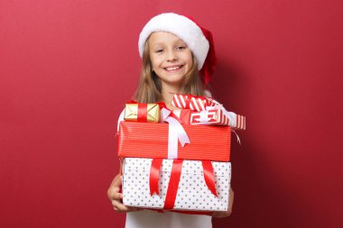 Renkli bir arka planda, Noel şapkası takmış, elinde hediyeler tutan sevimli, neşeli bir kız.