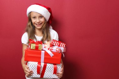 Renkli bir arka planda, Noel şapkası takmış, elinde hediyeler tutan sevimli, neşeli bir kız.