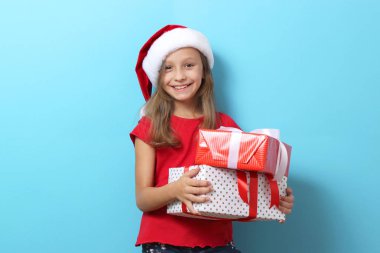 Renkli bir arka planda, Noel şapkası takmış, elinde hediyeler tutan sevimli, neşeli bir kız.