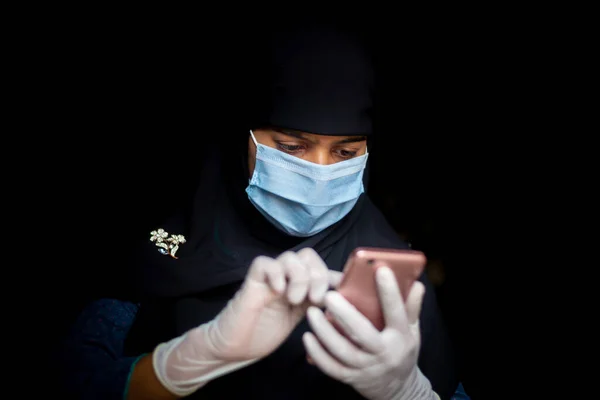 一个戴着安全面具和手套的穆斯林女孩正在他的智能手机上浏览验尸报告 为安全起见 戴蓝色面具的黑Hijab妇女 — 图库照片