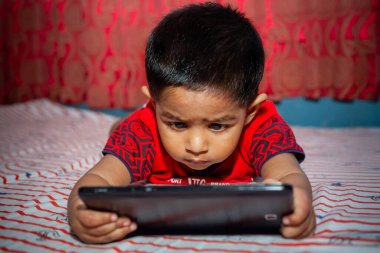 Tüm dikkatini yatağına vermiş, Tab akıllı telefonunu kullanarak çizgi film izleyen bir çocuk. Akıllı telefonla oynayan çocuklar. Cep telefonu ve internet bağımlılığı kavramı.