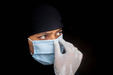 Müslüman kız Coronavirus koruması için cerrahi maske takıyor. Siyah tesettürlü kadın güvenlik için mavi bir maske takıyor. Koyu arkaplan görünümleri.