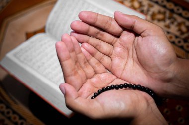 Genç Müslüman kadın Tasbeeh 'le namaz kılıyor. Kutsal Kur 'an arka planda, kapalı mekanda. Ellere odaklan..
