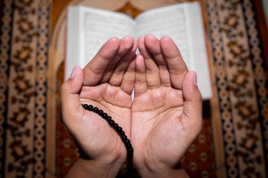Genç Müslüman kadın Tasbeeh 'le namaz kılıyor. Kutsal Kur 'an arka planda, kapalı mekanda. Ellere odaklan..