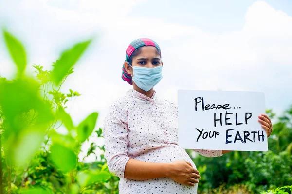 A young pregnant woman standing in green nature wearing a medical mask is calling for saving the earth from the coronavirus epidemic. A placard message to \