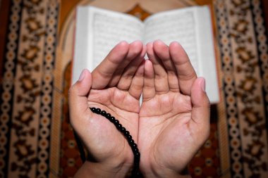 Genç Müslüman kadın Tasbeeh 'le namaz kılıyor. Kutsal Kur 'an arka planda, kapalı mekanda. Ellere odaklan..