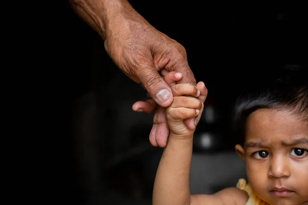 Bir erkek bebek kendinden emin bir şekilde yaşlı bir adamın parmağını tutarak etrafta dolaşıyor. Aile, Nesil, Destek ve İnsan Konsepti. Koyu arkaplan.