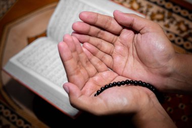 Genç Müslüman kadın Tasbeeh 'le namaz kılıyor. Kutsal Kur 'an arka planda, kapalı mekanda. Ellere odaklan..