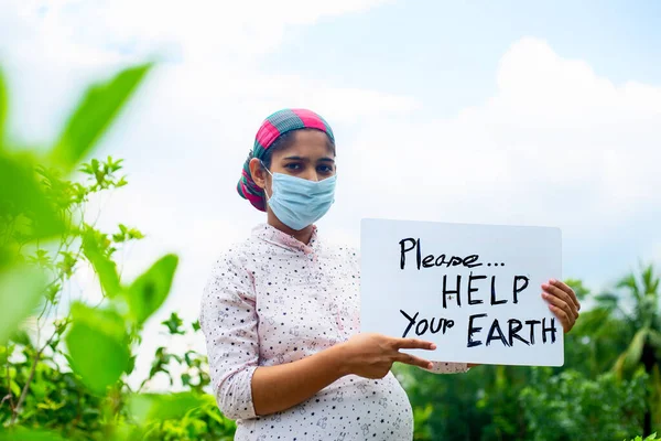 A young pregnant woman standing in green nature wearing a medical mask is calling for saving the earth from the coronavirus epidemic. A placard message to 