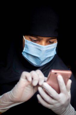 Güvenlik maskesi ve eldiven takan Müslüman bir kız Coronavirus haberleri için telefonuna bakıyor. Koronavirüs güvenliği için mavi maske takan siyah tesettürlü kadın..